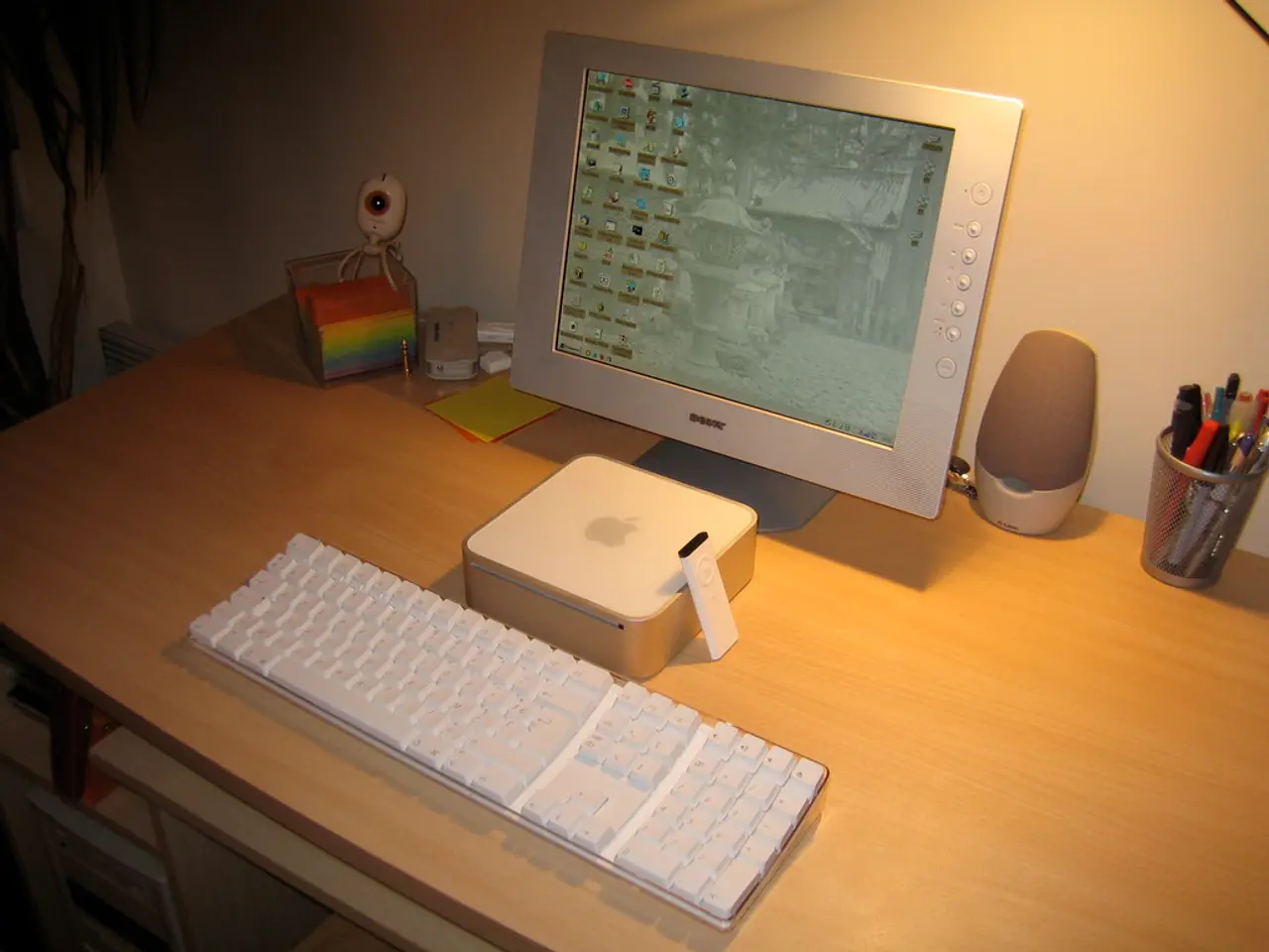 In this image on a table there is a monitor, keyboard, remote, speaker, a pen stand with pens, and...