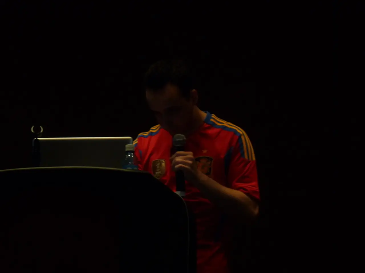 This image consists of a man wearing a red T-shirt is talking in a mic. In front of him, there is a...