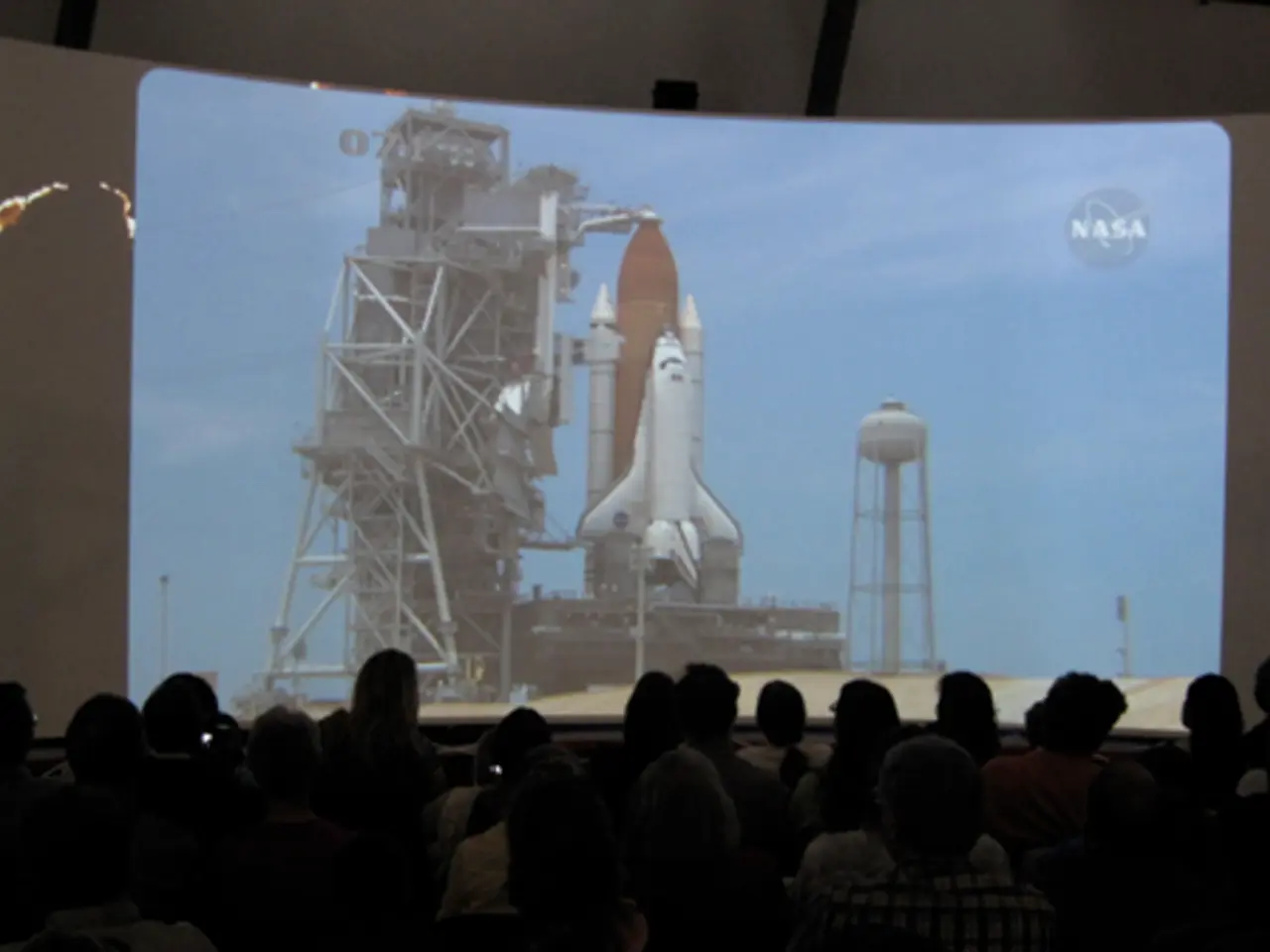 In this image, we can see a crowd in front of the screen. On the screen, there is a rocket...