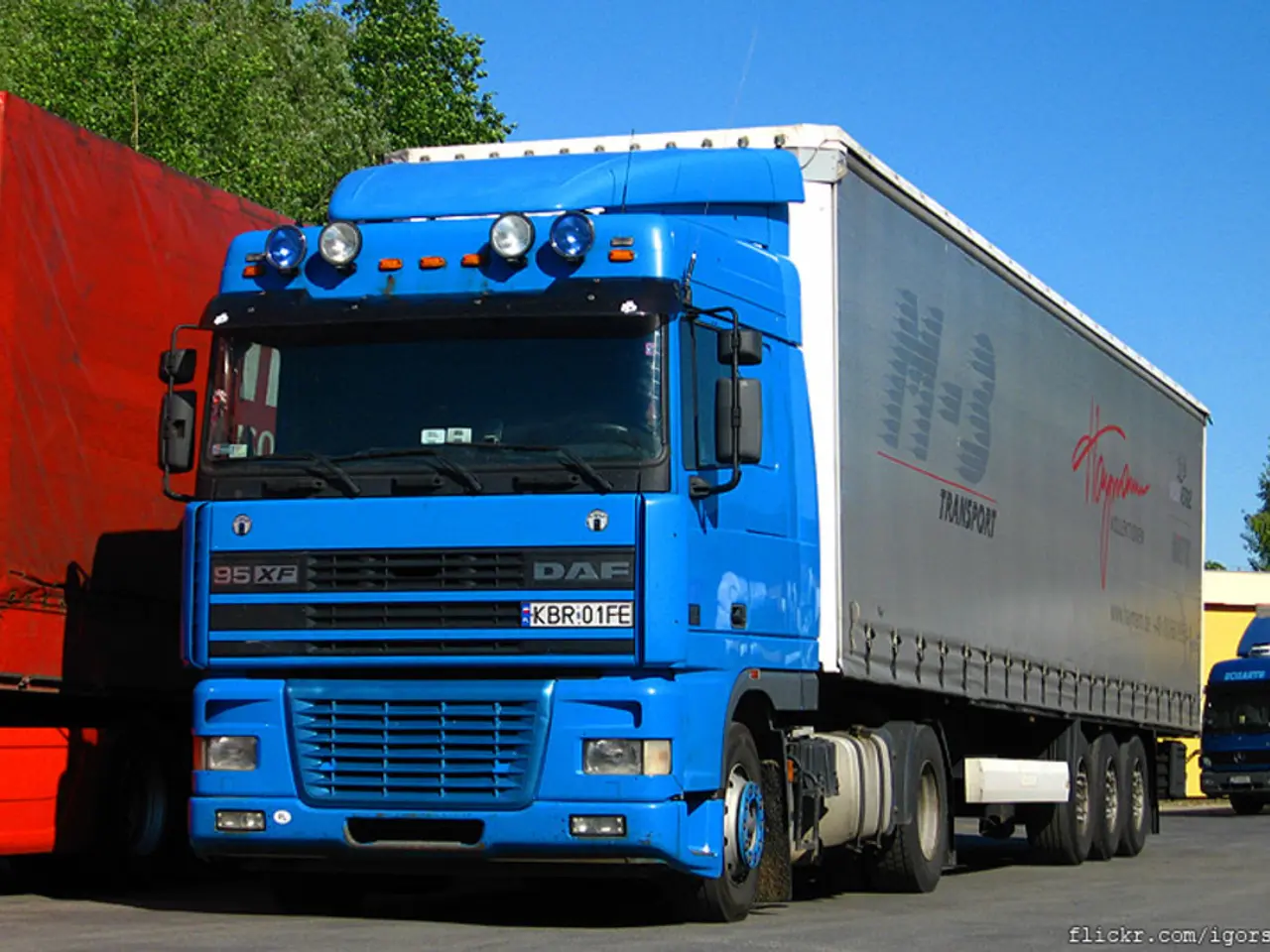 In this image I can see few blue colour trucks, a red colour thing, few trees, the sky and on this...