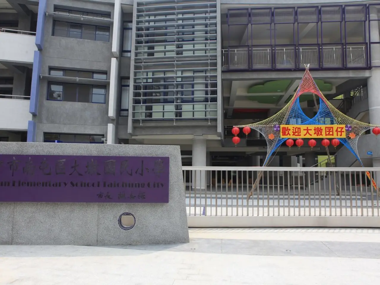 This picture shows a building and we see a name board on the wall. We see metal fence and few...