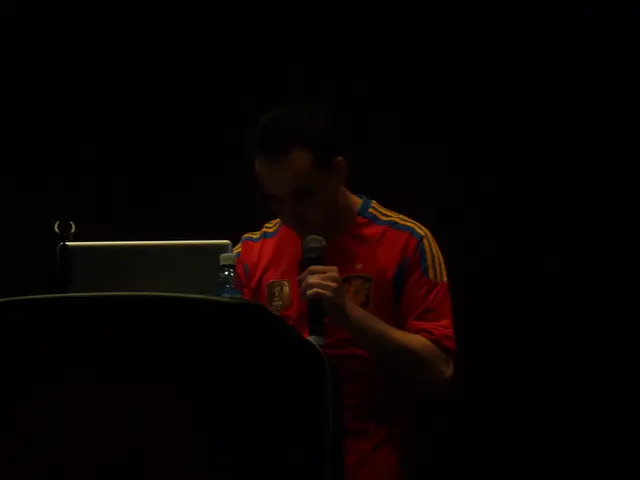This image consists of a man wearing a red T-shirt is talking in a mic. In front of him, there is a...