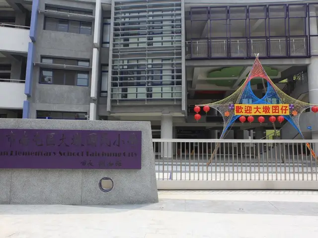 This picture shows a building and we see a name board on the wall. We see metal fence and few...