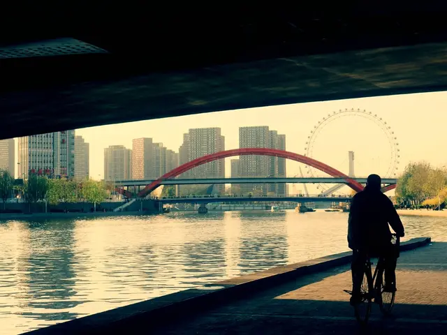 A man is riding a bicycle passing under a bridge by the side of a river. There is another bridge at...